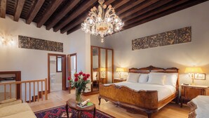 Une chambre spacieuse avec un grand lit, des poutres apparentes en bois au plafond, un lustre et un tapis rouge à motifs.