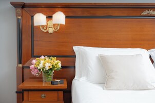 Cama bem arrumada com lençóis brancos, mesa de cabeceira de madeira com um vaso de flores e luminária fixada na parede.