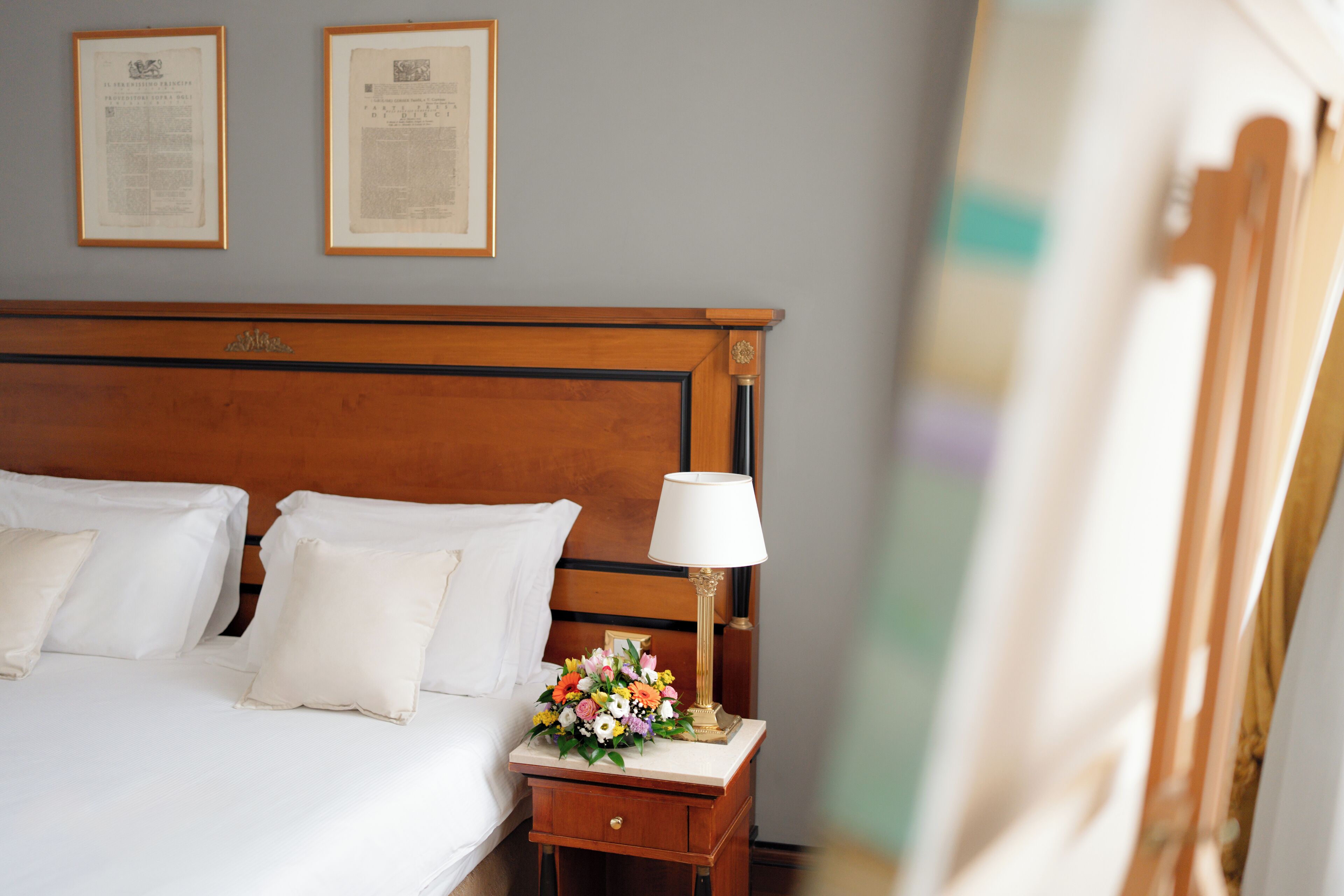 Ein ordentlich bezogenes Bett mit weißen Leinen und Kissen, ein Nachttisch mit Lampe und einer Blumenkomposition sowie zwei gerahmte Zertifikate an der Wand.