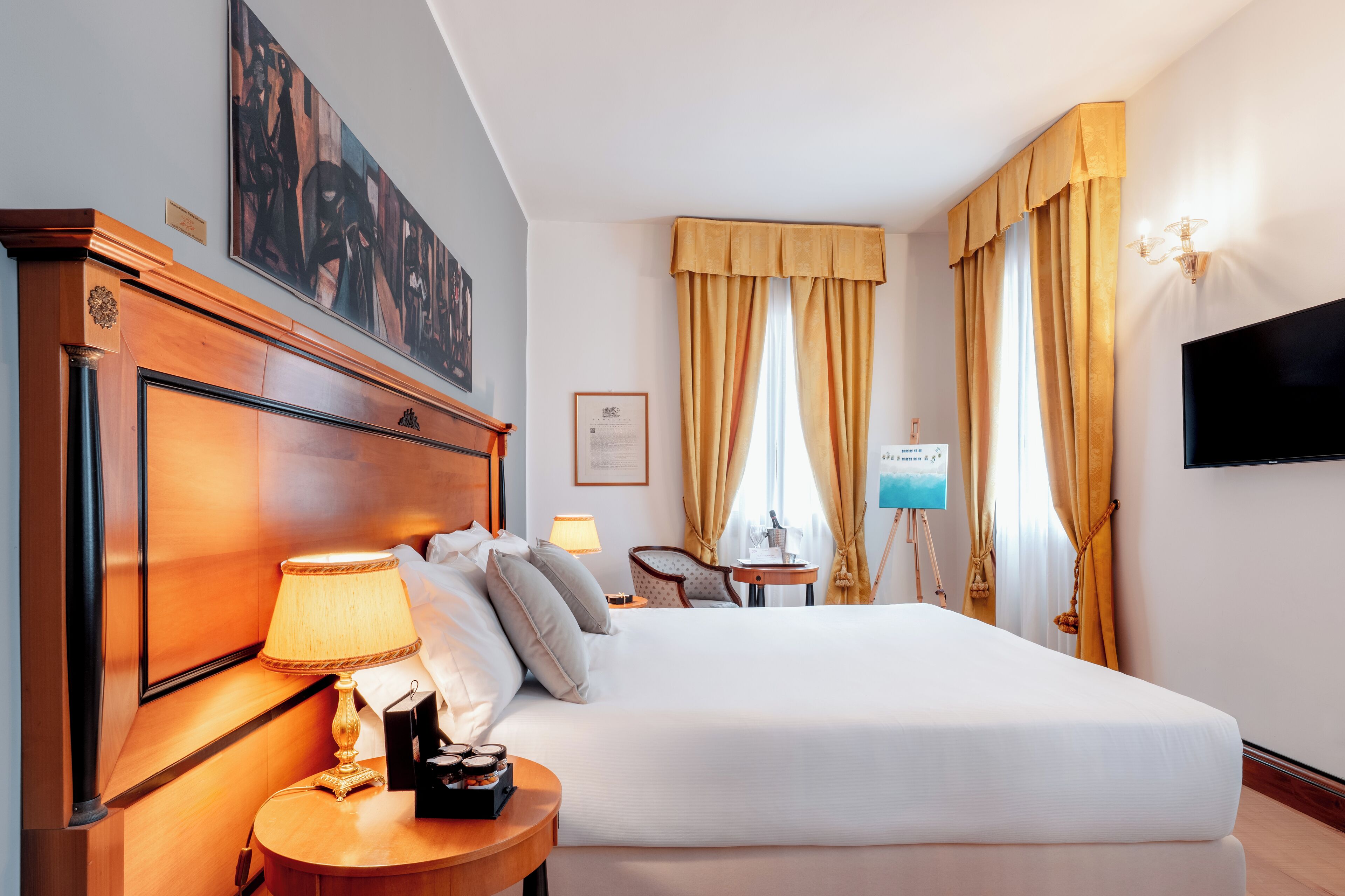Ein Hotelzimmer mit einem großen Bett, einem hölzernen Kopfteil, einem Nachttisch mit Lampe, einem kleinen Tisch mit einem Gemälde, einem Sessel und einem an der Wand montierten Fernseher.