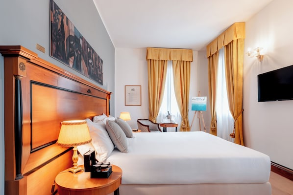 A hotel room with a large bed, a wooden headboard, a bedside table with a lamp, a small table with a painting, a chair, and a TV mounted on the wall. - All’Angelo Art Hotel (Venice)
