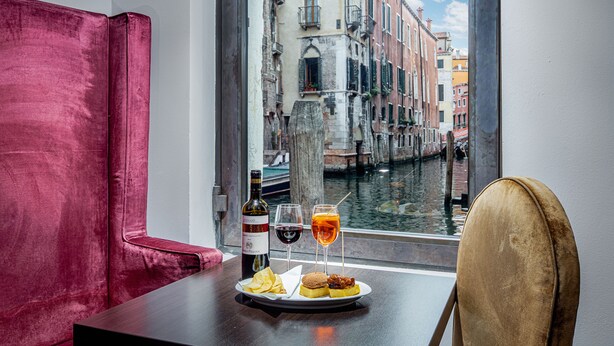 Une table avec une bouteille de vin, un verre de vin rouge, un cocktail et des amuse-gueules, avec vue sur un canal et des bâtiments, le tout visible à travers une fenêtre.
