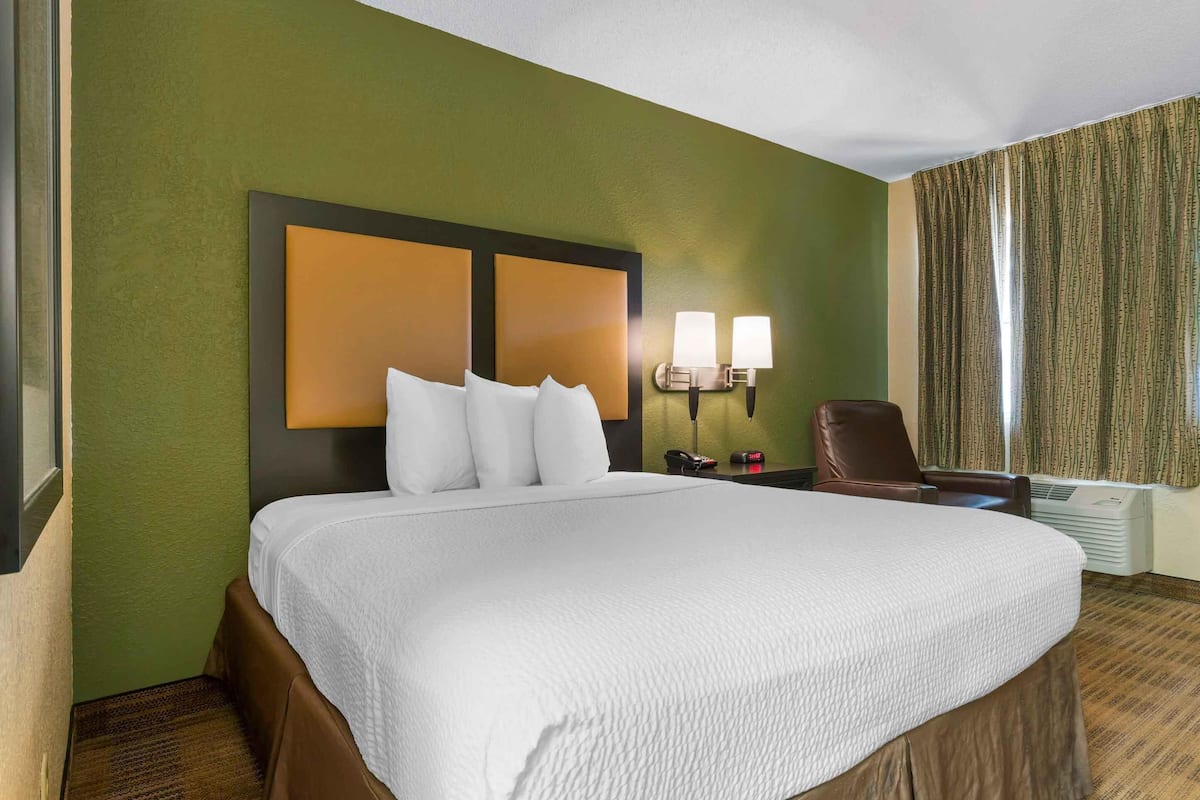 Ein Hotelzimmer mit einem großen Bett, zwei Nachttischlampen, einem Sessel und einem Fenster mit Vorhängen.