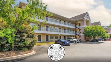 A two-story motel with a parking lot, a blue car, and a tree in the foreground.