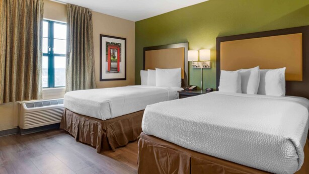 Habitación de hotel con dos camas, una ventana con cortinas, una lámpara de pared y un cuadro enmarcado.