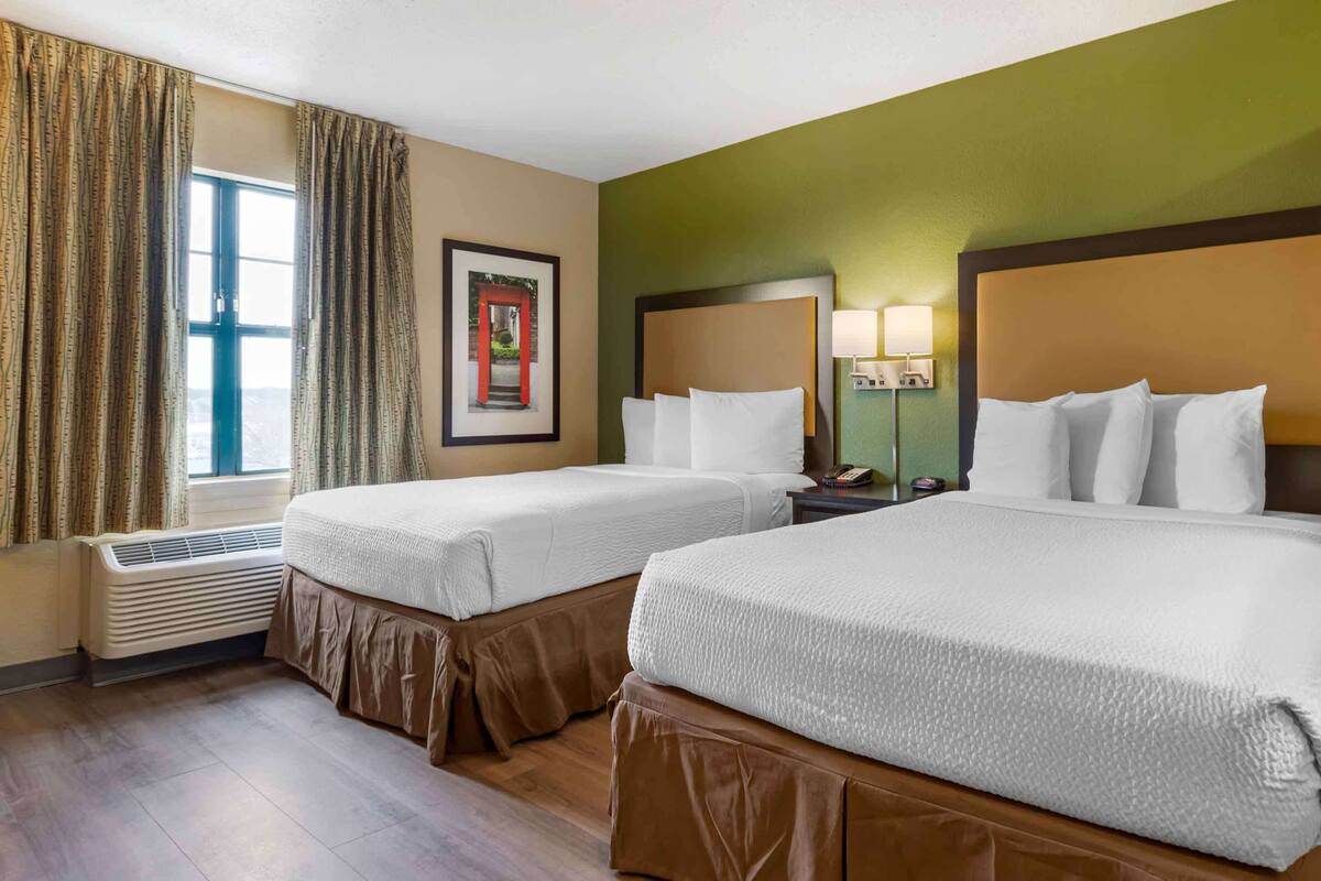 A hotel room with two beds, a window with curtains, a wall-mounted lamp, and a framed picture.