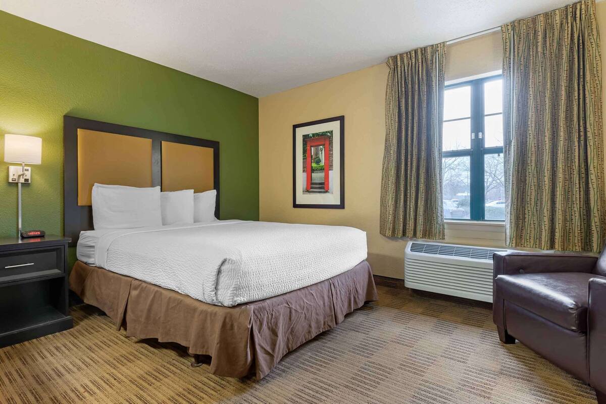 A hotel room with a large bed, a nightstand, a chair, a framed picture on the wall, and a window with curtains.