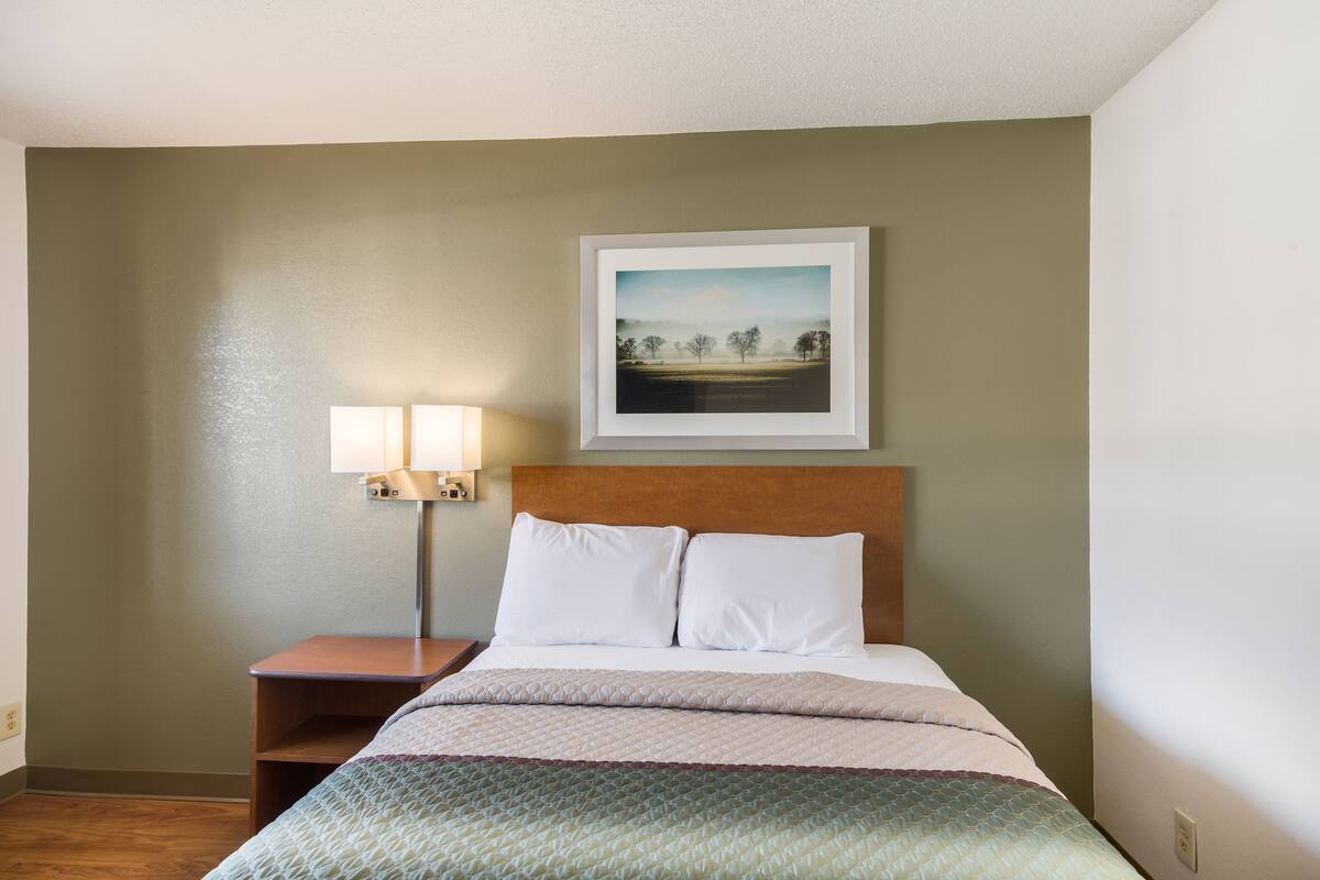 Ein ordentlich eingerichtetes Hotelzimmer mit einem Bett, einem Nachttisch mit Lampe und einem gerahmten Bild an der Wand.