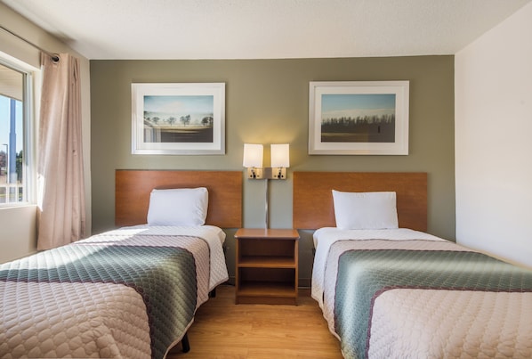 Une chambre d’hôtel avec deux lits, une table de chevet, deux lampes fixées au mur et deux tableaux encadrés accrochés au mur.