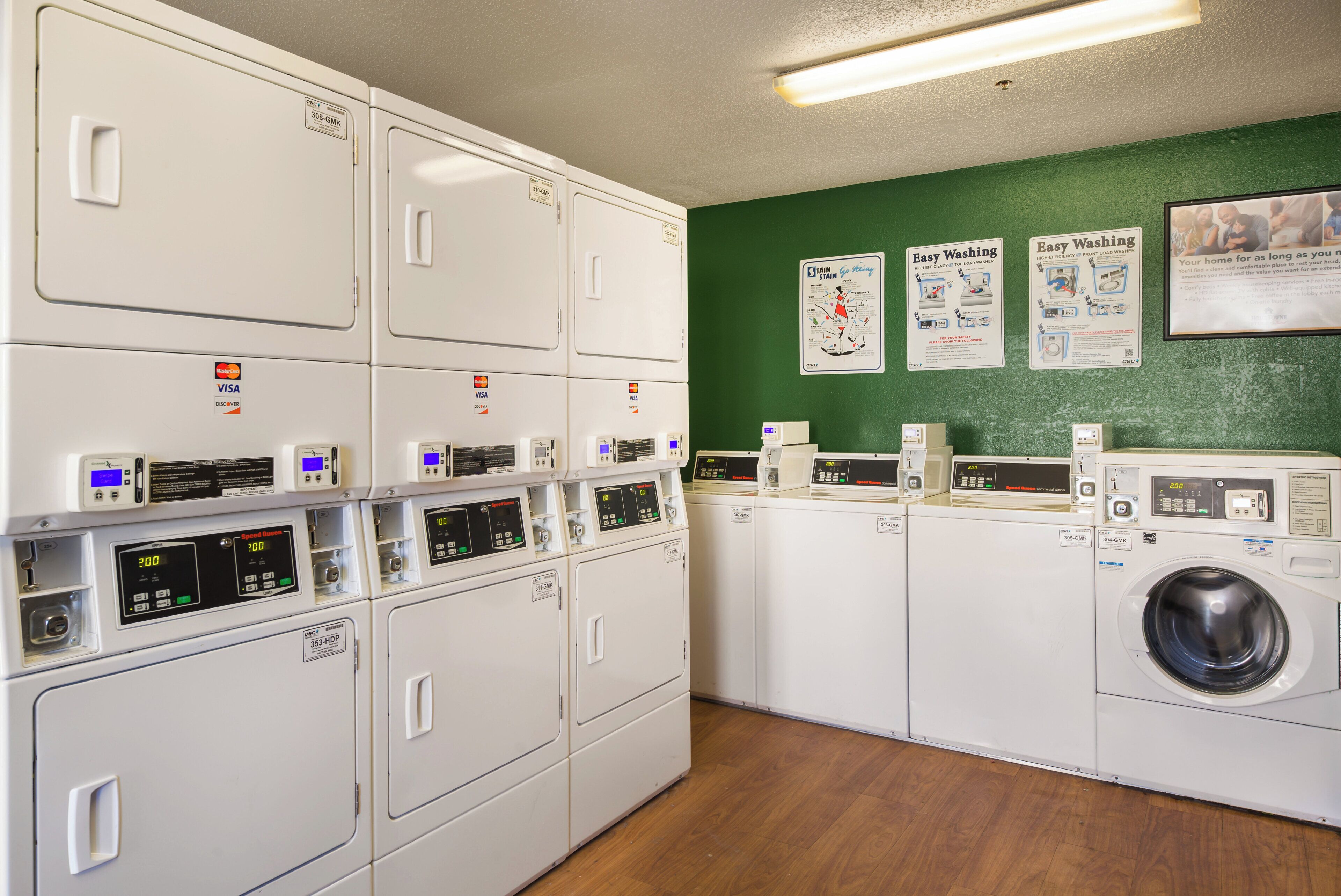A laundry facility with multiple washing machines and dryers, a green wall with informational posters, and a wooden floor.
