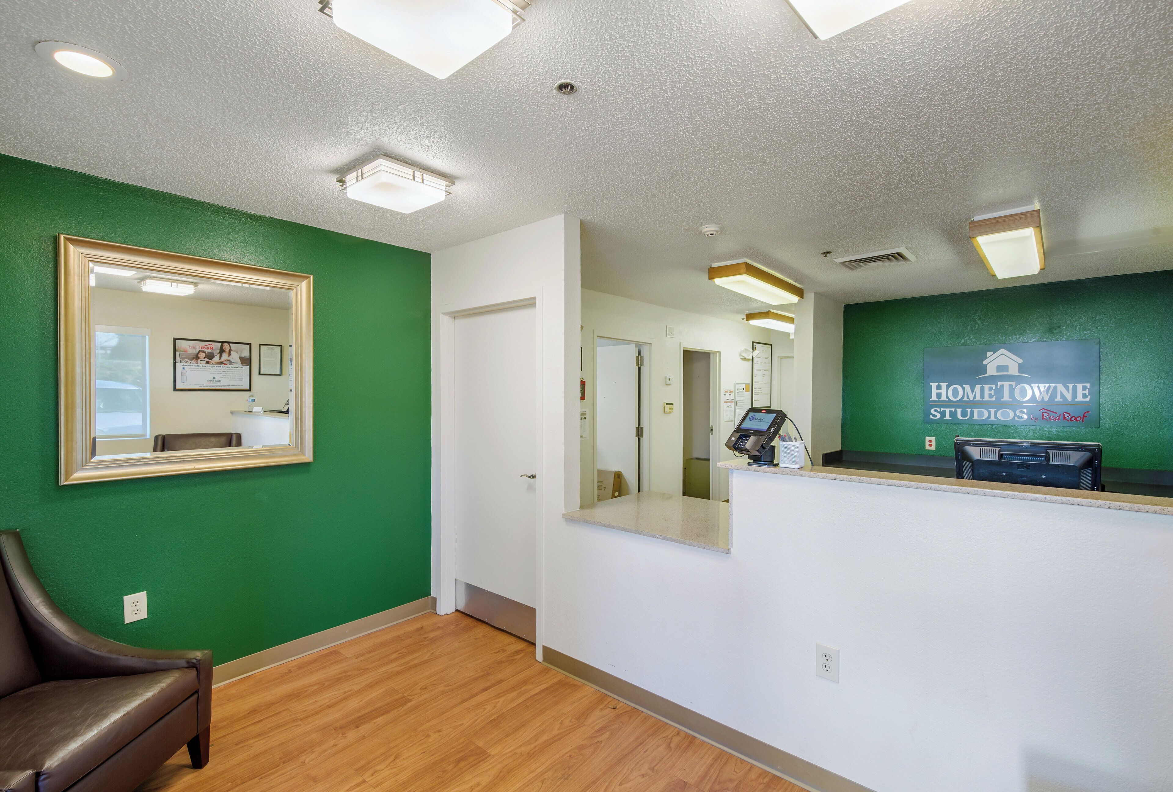 A reception area with a green wall, a framed mirror, a leather chair, and a reception desk with a computer and a sign reading 'HomeTowne Studios by Red Roof'.