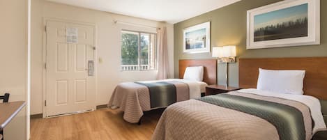 Habitación de hotel con dos camas, un escritorio, una silla, un ventanal con cortinas, una puerta y un cuadro en la pared.