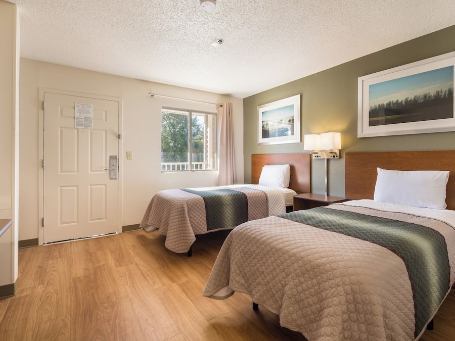 Habitación de hotel con dos camas, un escritorio, una silla, un ventanal con cortinas, una puerta y un cuadro en la pared.