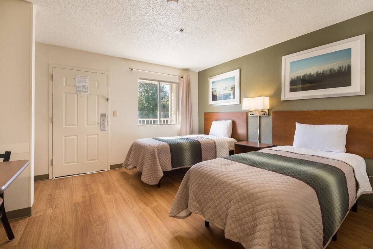 Ein Hotelzimmer mit zwei Betten, einem Fenster, einer Tür, einem gerahmten Bild an der Wand und einem kleinen Tisch.