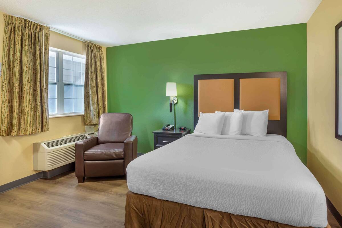 Ein Hotelzimmer mit einem großen Bett, einem Sessel, einem Nachttisch mit Lampe und einem Fenster mit Vorhängen.