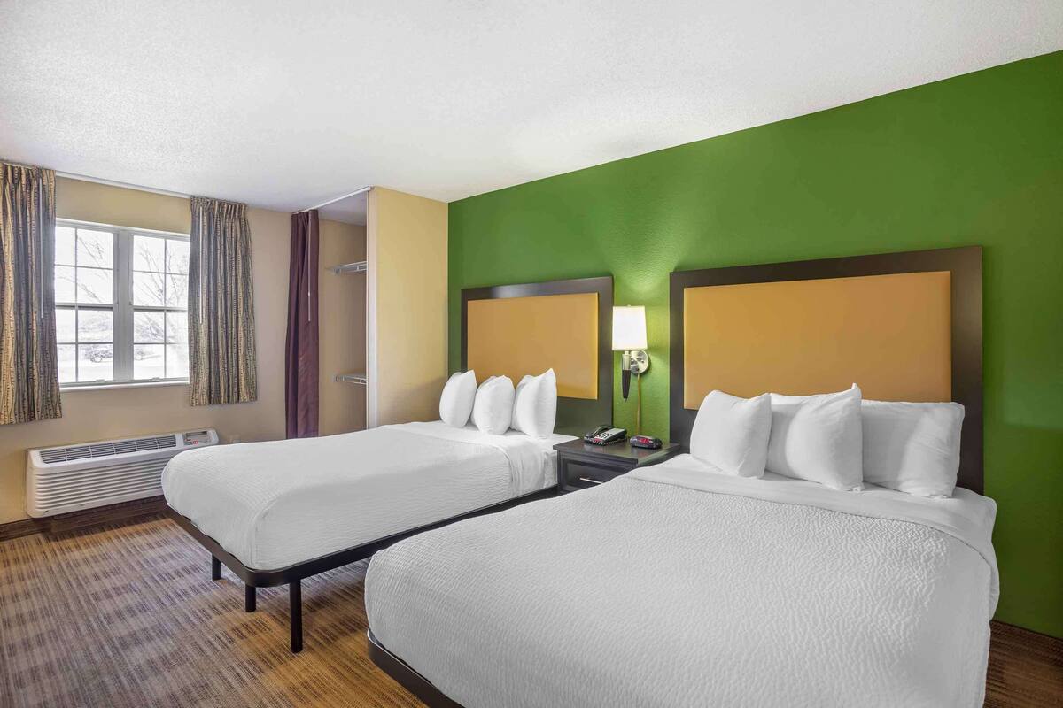 Ein Hotelzimmer mit zwei Betten, einem großen Fenster mit Vorhängen, einem Heizkörper und einer Wand in Grünton.