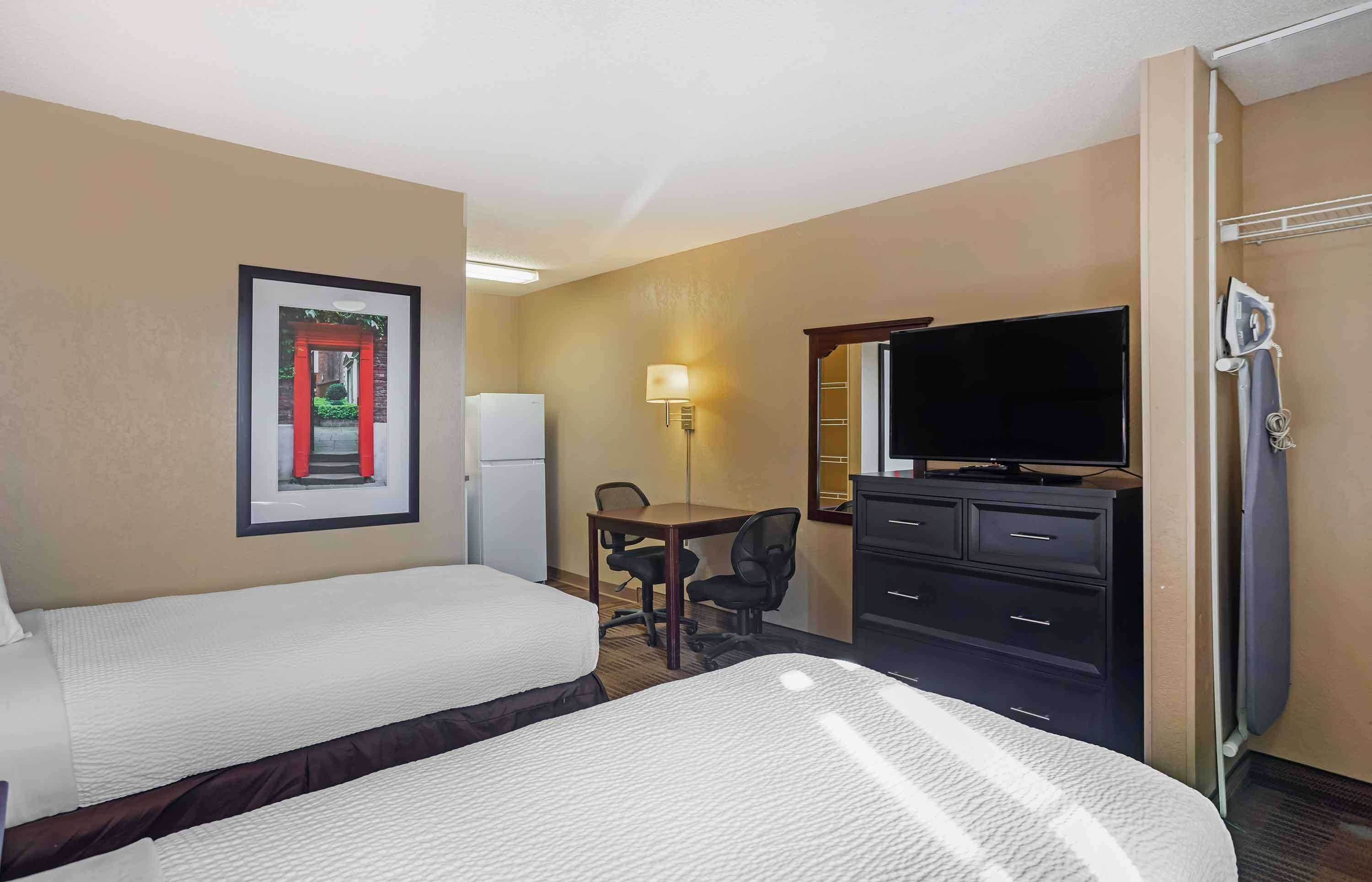 A hotel room with two beds, a desk with a chair, a television, a refrigerator, and a wall-mounted ironing board.
