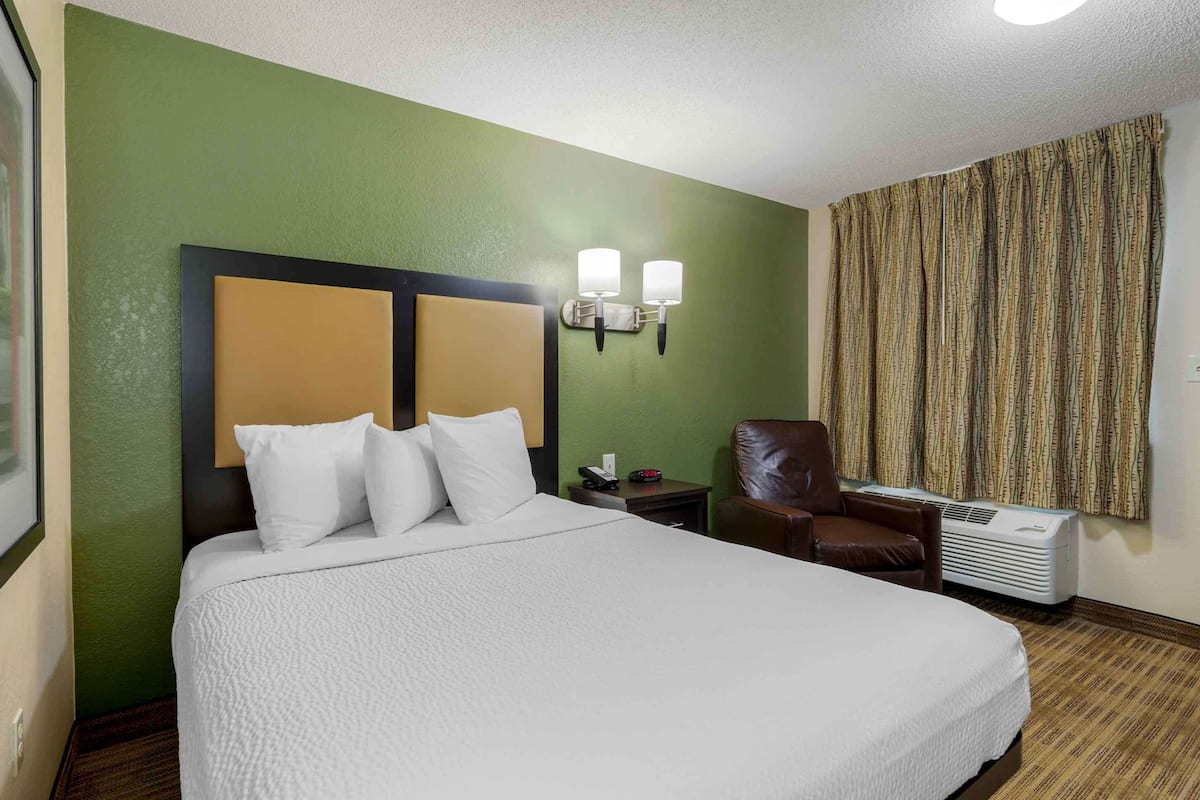 A hotel room with a large bed, a headboard, two bedside lamps, a chair, a nightstand, and an air conditioning unit.