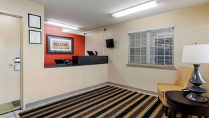 A hotel reception area with a reception desk, a 'WELCOME' sign, a TV, a window with blinds, a chair, and a lamp. - Extended Stay America Select Suites - Chattanooga - Airport (Chattanooga)