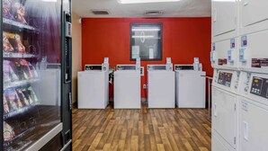 A laundry room with three washing machines, a vending machine, and a red wall. - Extended Stay America Select Suites - Chattanooga - Airport (Chattanooga)