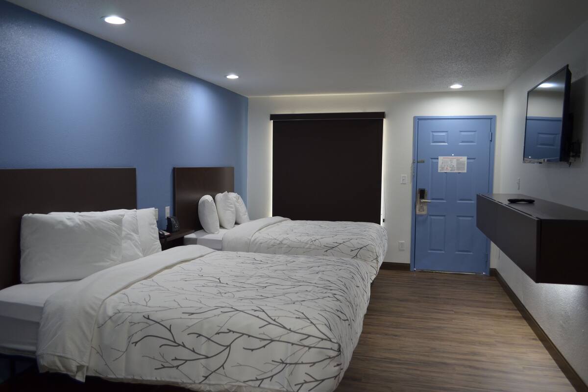 Quarto de hotel com duas camas, uma escrivaninha, uma cadeira, uma porta azul e um espelho na parede.