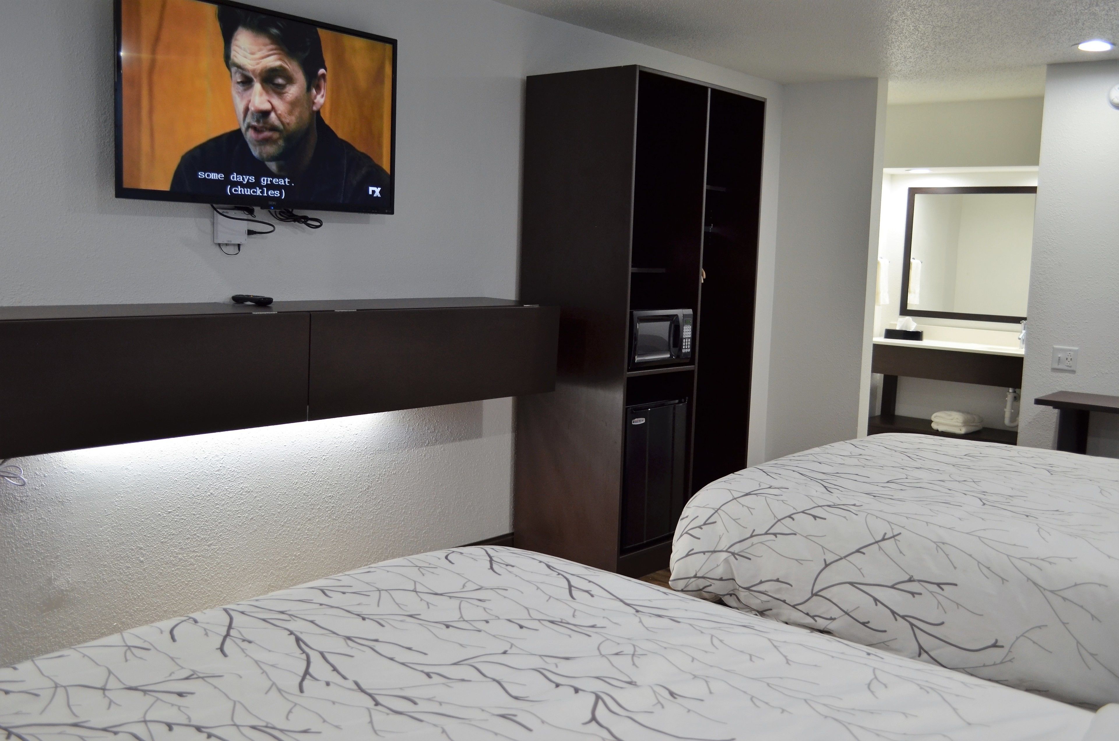 A hotel room with two beds, a TV mounted on the wall, a dark wardrobe, a microwave, and a small table with a mirror.
