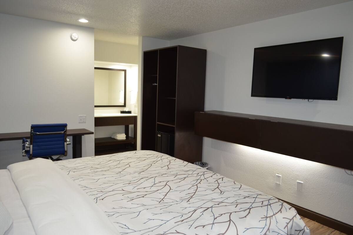 Quarto de hotel com uma cama, escrivaninha, cadeira, guarda-roupa, televisão e espelho.