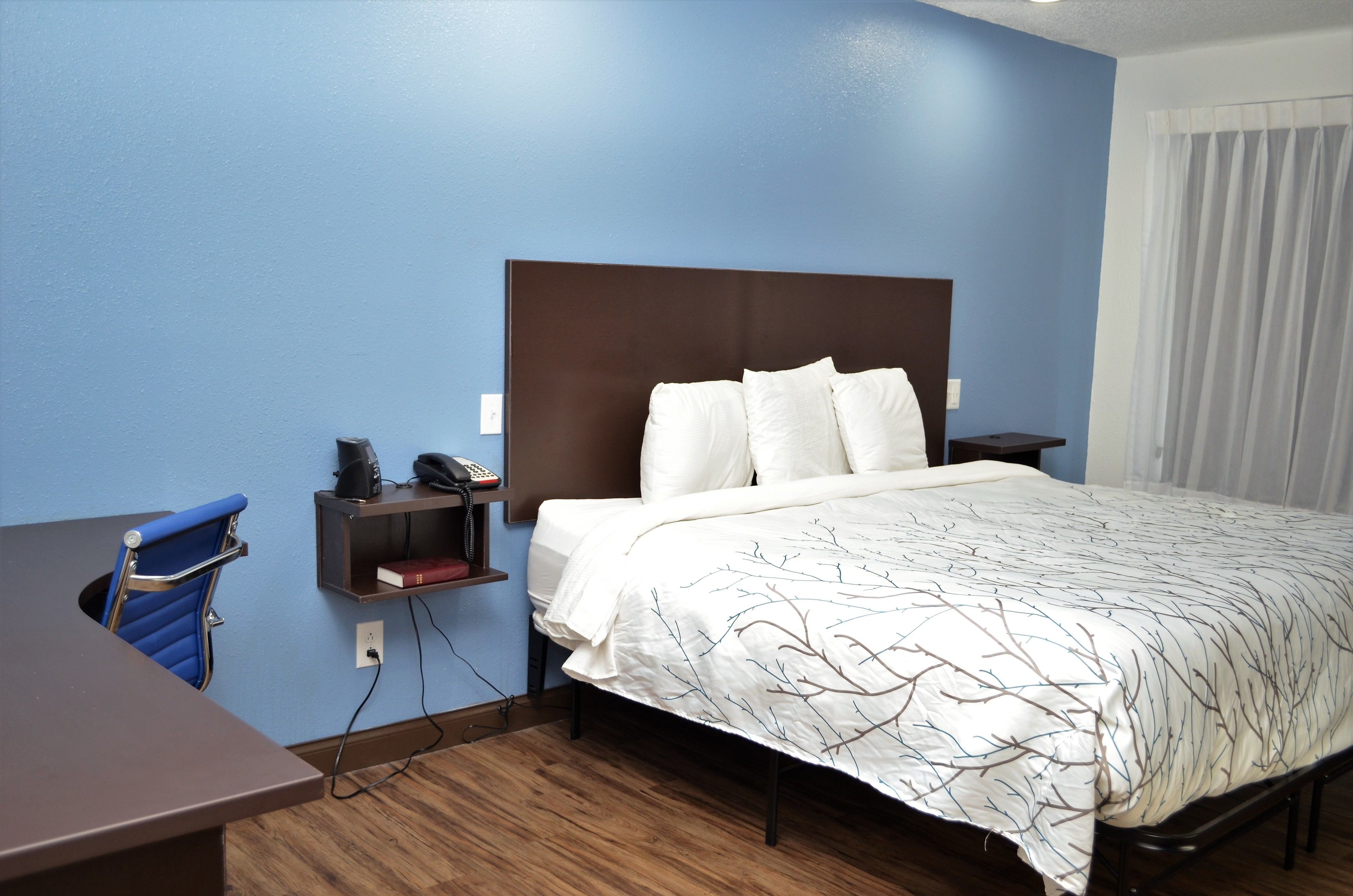 Quarto de hotel com cama, escrivaninha, cadeira e mesa de cabeceira.