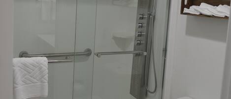 A modern bathroom with a glass-enclosed shower, a white toilet, and a wooden shelf with towels.