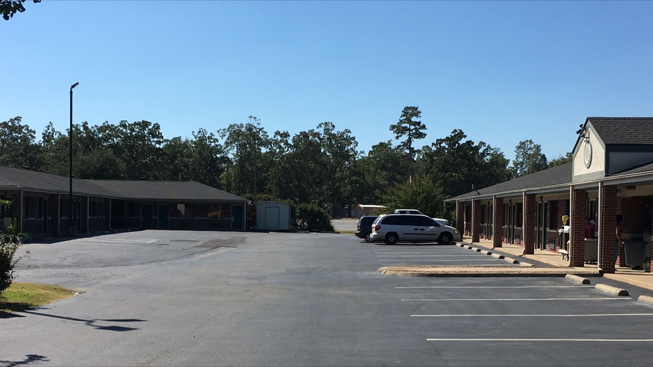 A parking lot with a row of single-story buildings, a white car parked in the lot, and trees in the background.
