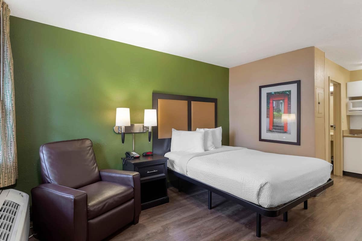 Habitación de hotel con cama, sillón, mesita de noche, lámpara, cuadro enmarcado y kitchenette.