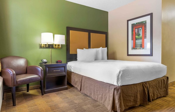 Une chambre d’hôtel avec un lit, une chaise, une table de chevet avec une lampe et un tableau encadré au mur.