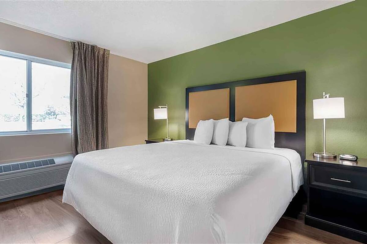 A hotel room with a large bed, two bedside tables with lamps, a window with curtains, and a green wall.