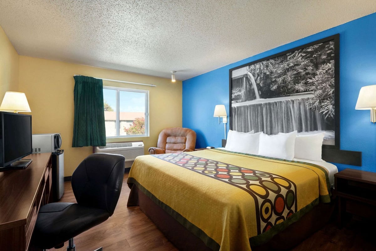 A hotel room with a bed, a desk with a computer, a chair, a window with curtains, and a wall-mounted TV.