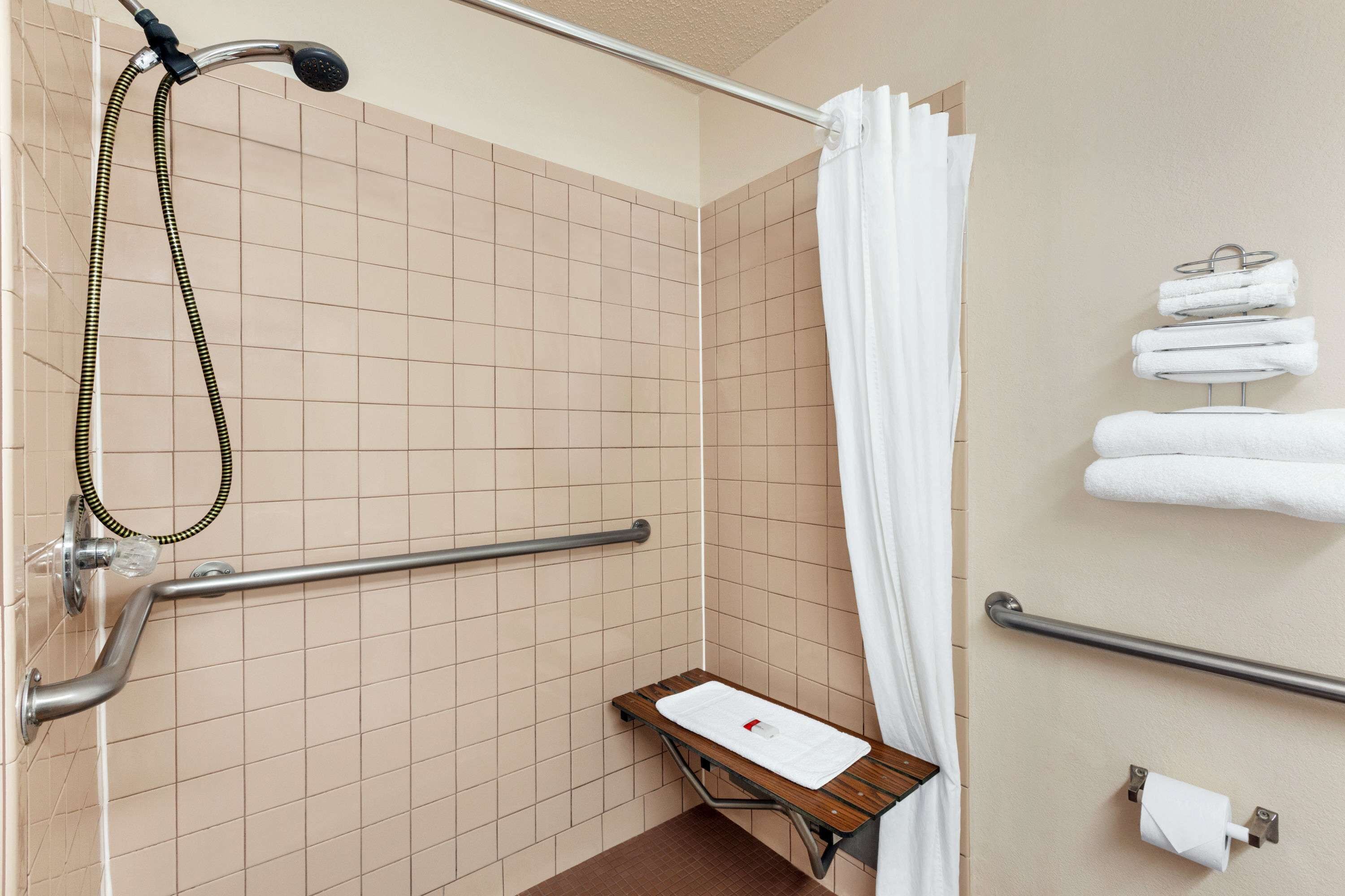 A bathroom with beige tiled walls, a shower with a handheld hose, a grab bar, a wooden bench, and a towel rack with folded towels.