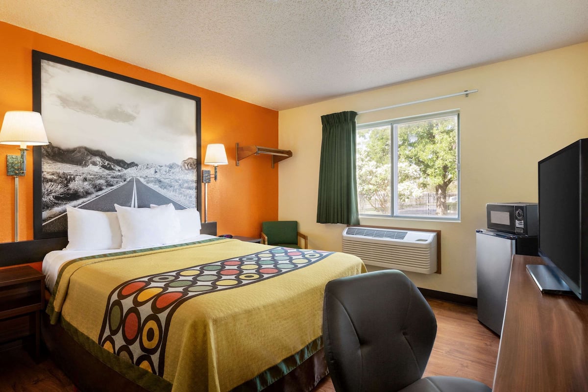 A hotel room with a bed, a desk, a television, a window with curtains, and a wall-mounted air conditioner.