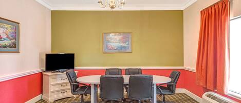 Une salle de conférence avec une table ronde, six chaises, un téléviseur à écran plat, un tableau accroché au mur et une fenêtre avec des rideaux.