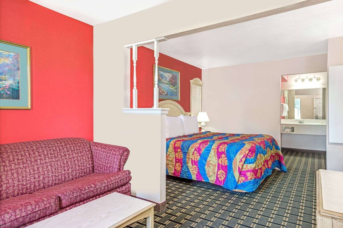 Ein Hotelzimmer mit roter Wand, einem Bett mit farbenfroher Steppdecke, einer Couch, einem kleinen Tisch und einem Spiegel.
