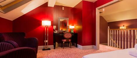 Habitación de hotel con techo inclinado, cama, escritorio con silla, espejo y pared roja.