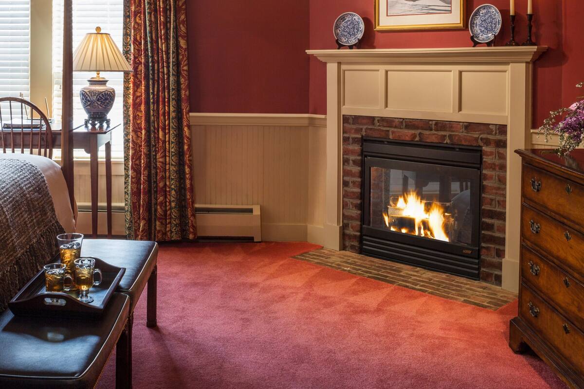 A hotel room with a four-poster bed, a fireplace, a wooden dresser, and a dining area with a table and chairs.