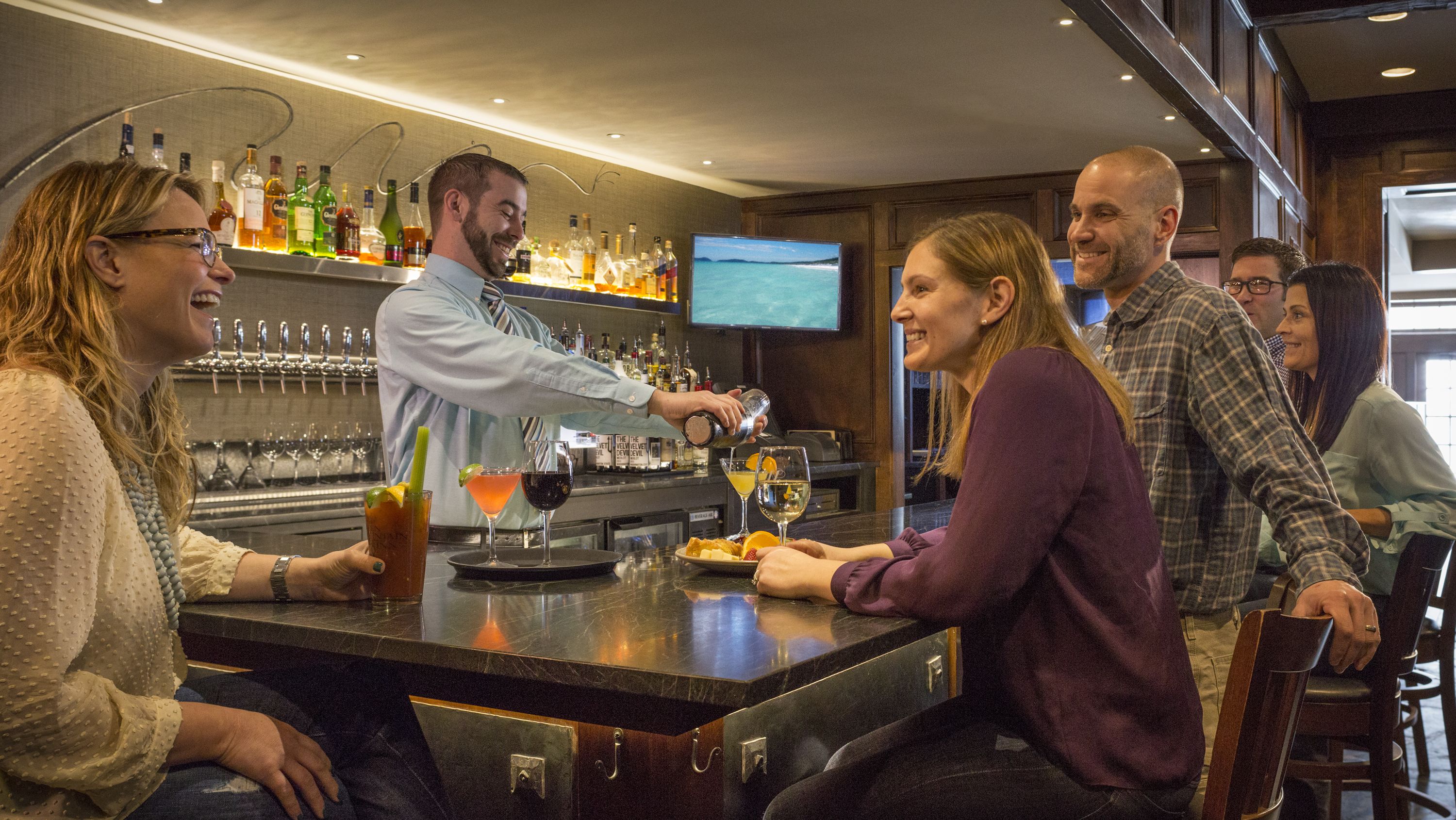 Zona de bar amb un barman preparant begudes, un televisor fixat a la paret i una barra plena de garrafes i gots.