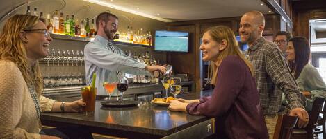 A bar area with a bartender preparing drinks, a television mounted on the wall, and a bar counter with various bottles and glasses.