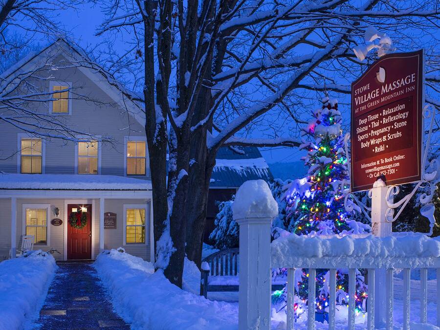 A snow-covered house with a lit front door, a Christmas tree with lights, and a sign for Stowe Village Massage.