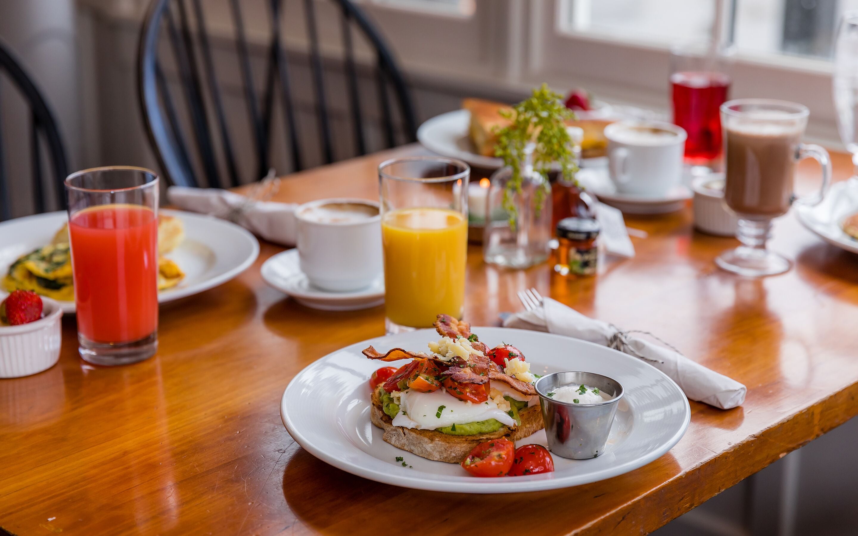 Colazione servita con pane tostato farcito con uova, pomodori e bacon, insieme a un bicchiere di succo d'arancia e una tazza di caffè.