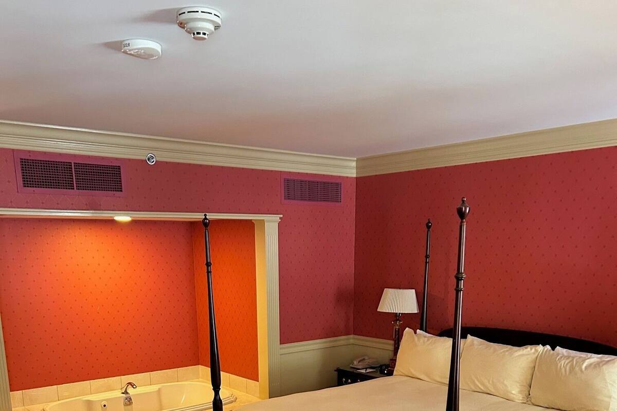 A four-poster bed with a white bedspread, a large window, and a bathroom with a bathtub and vanity.