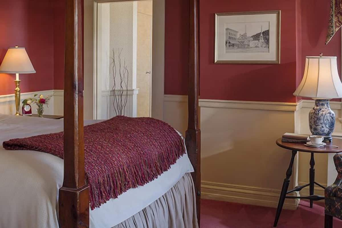 A four-poster bed with a red throw blanket, a bedside table with a lamp, and a chair.
