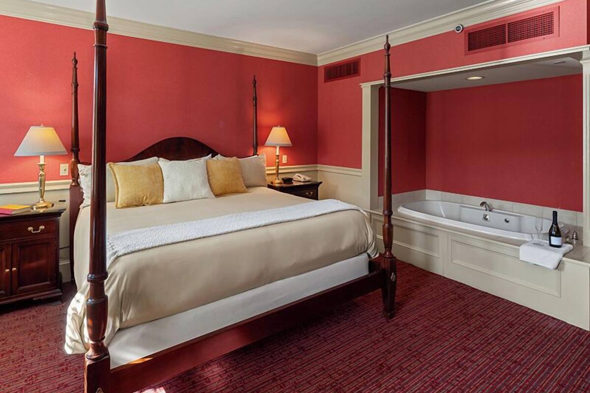 A four-poster bed with a wooden frame, a large mirror, and a bathtub in a room with red walls and a red carpet.