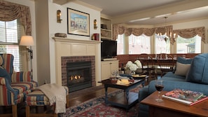 A living room with a fireplace, striped armchairs, a sofa, a coffee table with a book and wine glass, and a television above the fireplace.