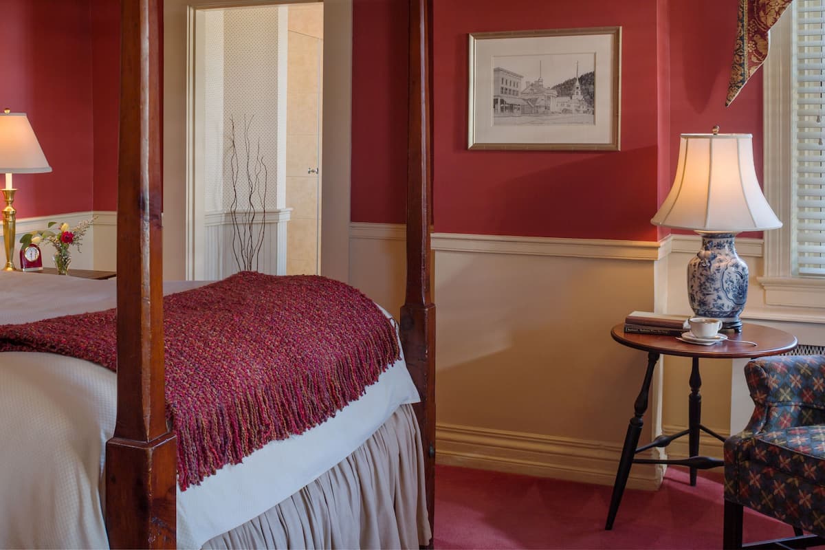 A four-poster bed with a red throw blanket, a small table with a lamp, and a chair with a patterned cushion.
