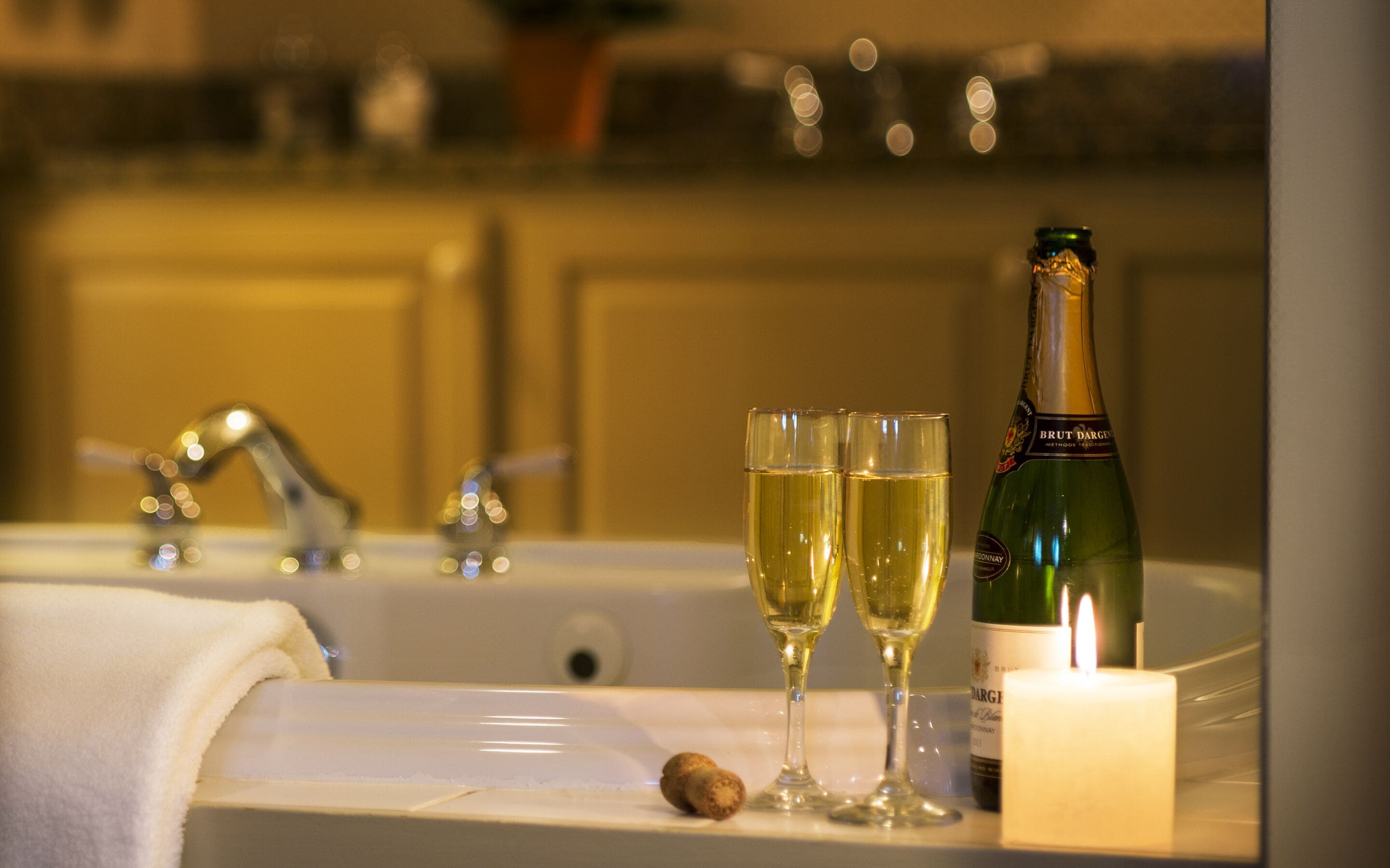 Un baño con una bañera, dos copas de champán, una botella de vino Brut Dargies y una vela encendida.